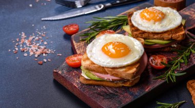 Fresh, delicious sandwiches with fried egg, ham, butter, avocado and sesame seeds on a wooden cutting board. Delicious healthy food