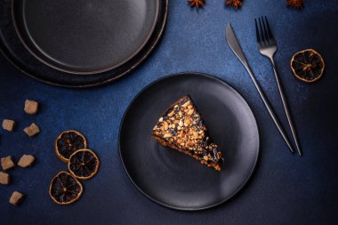 A piece of fresh delicious cake with nuts and chocolate on a black plate against a dark blue background. Desserts and sweets to the dinner table