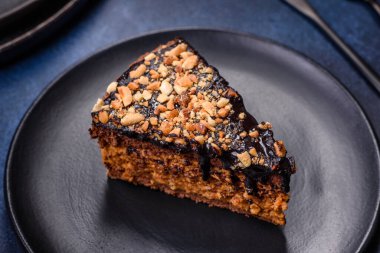 A piece of fresh delicious cake with nuts and chocolate on a black plate against a dark blue background. Desserts and sweets to the dinner table