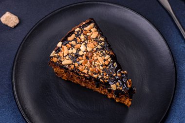 A piece of fresh delicious cake with nuts and chocolate on a black plate against a dark blue background. Desserts and sweets to the dinner table