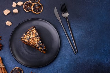 A piece of fresh delicious cake with nuts and chocolate on a black plate against a dark blue background. Desserts and sweets to the dinner table