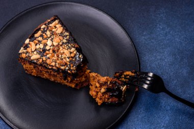 A piece of fresh delicious cake with nuts and chocolate on a black plate against a dark blue background. Desserts and sweets to the dinner table