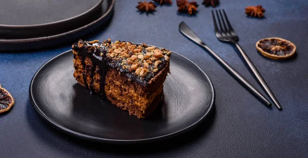 A piece of fresh delicious cake with nuts and chocolate on a black plate against a dark blue background. Desserts and sweets to the dinner table