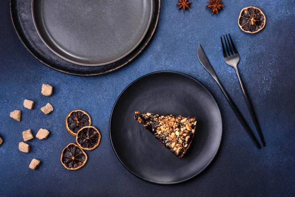 A piece of fresh delicious cake with nuts and chocolate on a black plate against a dark blue background. Desserts and sweets to the dinner table