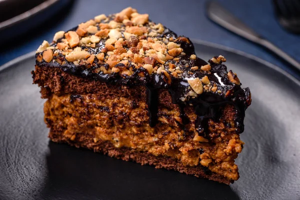 A piece of fresh delicious cake with nuts and chocolate on a black plate against a dark blue background. Desserts and sweets to the dinner table