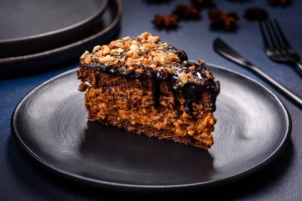 A piece of fresh delicious cake with nuts and chocolate on a black plate against a dark blue background. Desserts and sweets to the dinner table