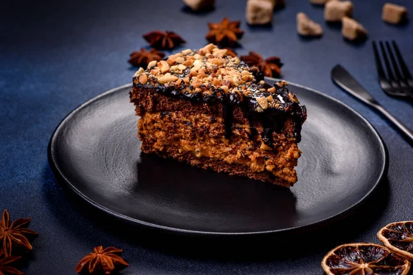 A piece of fresh delicious cake with nuts and chocolate on a black plate against a dark blue background. Desserts and sweets to the dinner table
