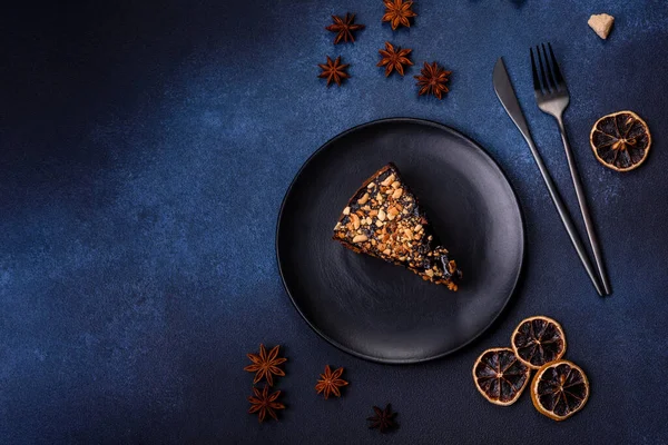 A piece of fresh delicious cake with nuts and chocolate on a black plate against a dark blue background. Desserts and sweets to the dinner table
