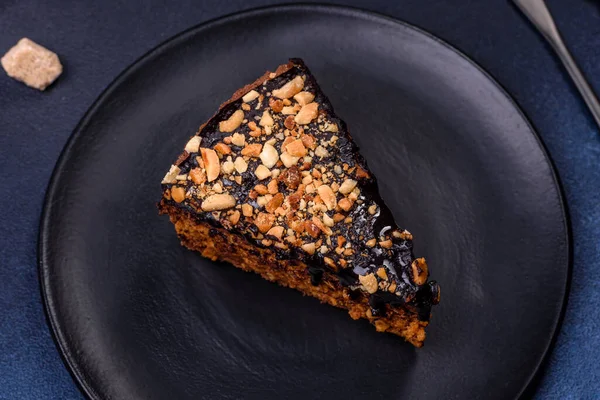 A piece of fresh delicious cake with nuts and chocolate on a black plate against a dark blue background. Desserts and sweets to the dinner table