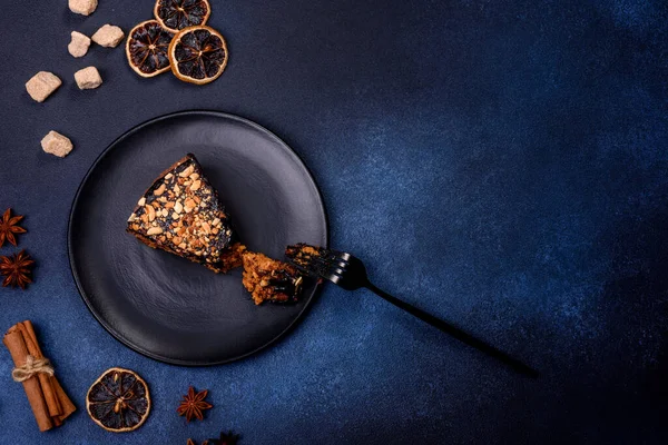 A piece of fresh delicious cake with nuts and chocolate on a black plate against a dark blue background. Desserts and sweets to the dinner table
