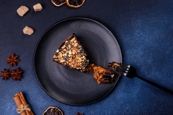 A piece of fresh delicious cake with nuts and chocolate on a black plate against a dark blue background. Desserts and sweets to the dinner table