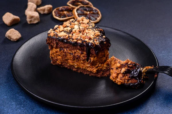 A piece of fresh delicious cake with nuts and chocolate on a black plate against a dark blue background. Desserts and sweets to the dinner table