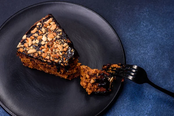 A piece of fresh delicious cake with nuts and chocolate on a black plate against a dark blue background. Desserts and sweets to the dinner table