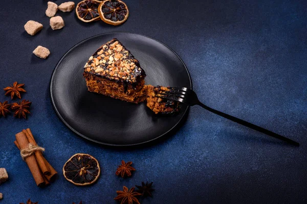A piece of fresh delicious cake with nuts and chocolate on a black plate against a dark blue background. Desserts and sweets to the dinner table