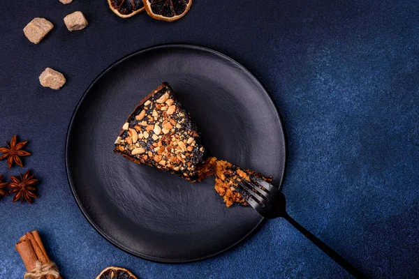 A piece of fresh delicious cake with nuts and chocolate on a black plate against a dark blue background. Desserts and sweets to the dinner table