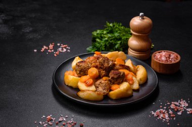 Beef meat and vegetables stew on a black plate with roasted potatoes. Dark background. Copy space