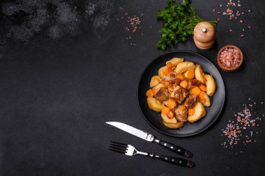 Beef meat and vegetables stew on a black plate with roasted potatoes. Dark background. Copy space