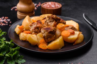 Beef meat and vegetables stew on a black plate with roasted potatoes. Dark background. Copy space