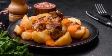 Beef meat and vegetables stew on a black plate with roasted potatoes. Dark background. Copy space
