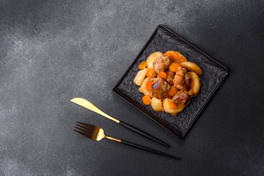 Beef meat and vegetables stew on a black plate with roasted potatoes. Dark background. Copy space
