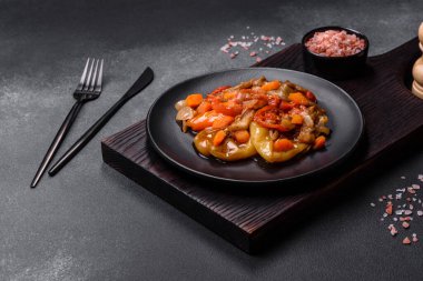 Baked vegetables, eggplant, peppers and carrots on a black plate on a gray concrete background. Vegetarian food