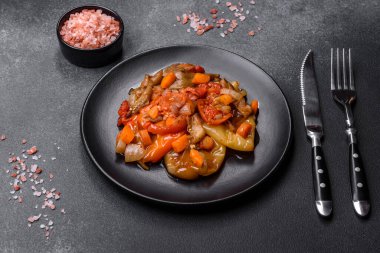 Baked vegetables, eggplant, peppers and carrots on a black plate on a gray concrete background. Vegetarian food