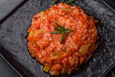 Boiled rice with peppers, spices, herbs and tomato sauce on a black slate plate on a grey concrete background
