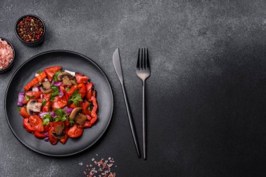 A salad of baked mushrooms, tomatoes, onions, parsley, spices and herbs on a black plate against a dark concrete background. Vegetarian cuisine dish