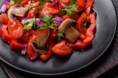 A salad of baked mushrooms, tomatoes, onions, parsley, spices and herbs on a black plate against a dark concrete background. Vegetarian cuisine dish