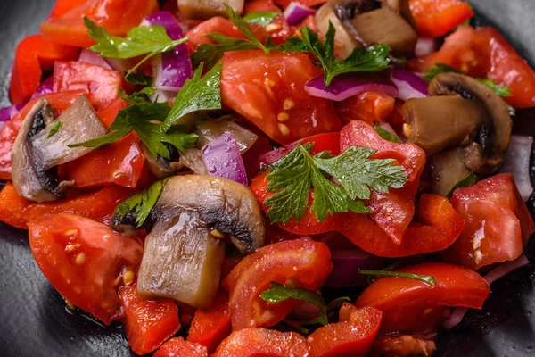 A salad of baked mushrooms, tomatoes, onions, parsley, spices and herbs on a black plate against a dark concrete background. Vegetarian cuisine dish