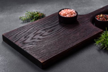 Wooden cutting board, salt, pepper, spices and herbs on a dark concrete background. Cooking delicious home-cooked food