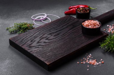 Wooden cutting board, salt, pepper, spices and herbs on a dark concrete background. Cooking delicious home-cooked food