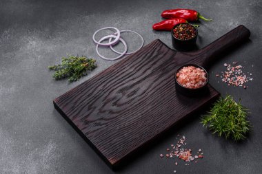 Wooden cutting board, salt, pepper, spices and herbs on a dark concrete background. Cooking delicious home-cooked food