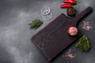 Wooden cutting board, salt, pepper, spices and herbs on a dark concrete background. Cooking delicious home-cooked food
