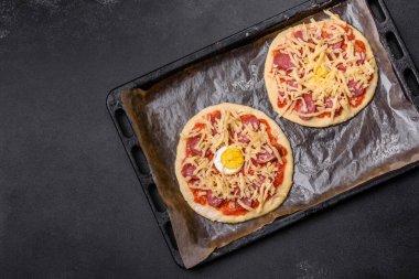 Homemade pizza with sausages, tomatoes, cheese, spices and herbs on a wooden cutting board against a dark concrete background