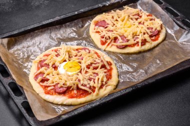 Homemade pizza with sausages, tomatoes, cheese, spices and herbs on a wooden cutting board against a dark concrete background