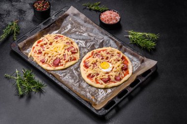 Homemade pizza with sausages, tomatoes, cheese, spices and herbs on a wooden cutting board against a dark concrete background