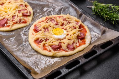 Homemade pizza with sausages, tomatoes, cheese, spices and herbs on a wooden cutting board against a dark concrete background