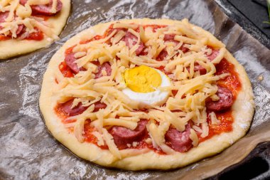 Homemade pizza with sausages, tomatoes, cheese, spices and herbs on a wooden cutting board against a dark concrete background