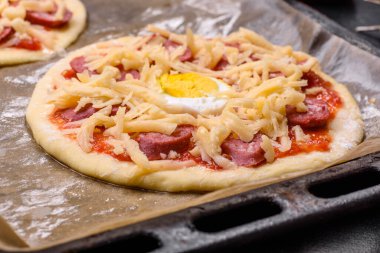 Homemade pizza with sausages, tomatoes, cheese, spices and herbs on a wooden cutting board against a dark concrete background