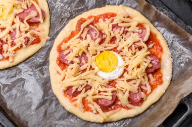 Homemade pizza with sausages, tomatoes, cheese, spices and herbs on a wooden cutting board against a dark concrete background