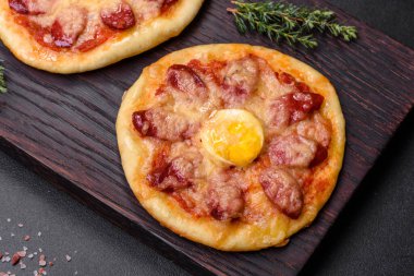 Homemade pizza with sausages, tomatoes, cheese, spices and herbs on a wooden cutting board against a dark concrete background