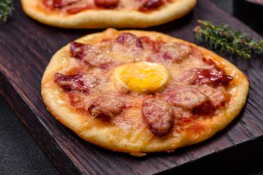 Homemade pizza with sausages, tomatoes, cheese, spices and herbs on a wooden cutting board against a dark concrete background