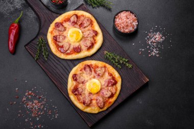 Homemade pizza with sausages, tomatoes, cheese, spices and herbs on a wooden cutting board against a dark concrete background