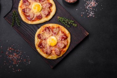 Homemade pizza with sausages, tomatoes, cheese, spices and herbs on a wooden cutting board against a dark concrete background