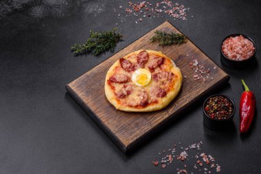 Homemade pizza with sausages, tomatoes, cheese, spices and herbs on a wooden cutting board against a dark concrete background