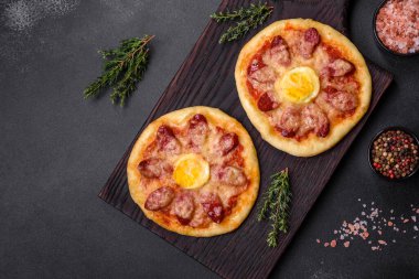 Homemade pizza with sausages, tomatoes, cheese, spices and herbs on a wooden cutting board against a dark concrete background