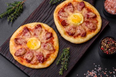Homemade pizza with sausages, tomatoes, cheese, spices and herbs on a wooden cutting board against a dark concrete background