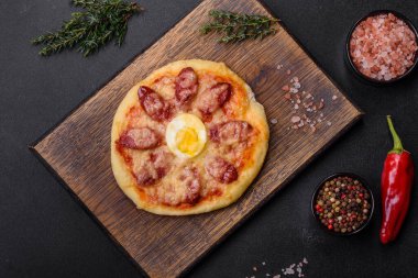 Homemade pizza with sausages, tomatoes, cheese, spices and herbs on a wooden cutting board against a dark concrete background