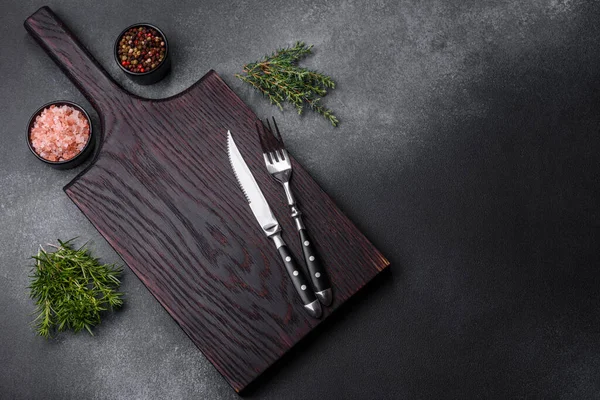 Wooden cutting board, salt, pepper, spices and herbs on a dark concrete background. Cooking delicious home-cooked food